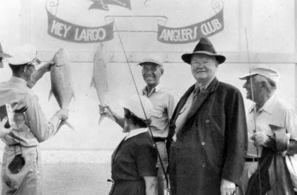 Black-and-white photograph of Herbert Hoover in a fedora and overcoat standing at the Key Largo Anglers Club beside two large fish hanging from the club's painted backdrop