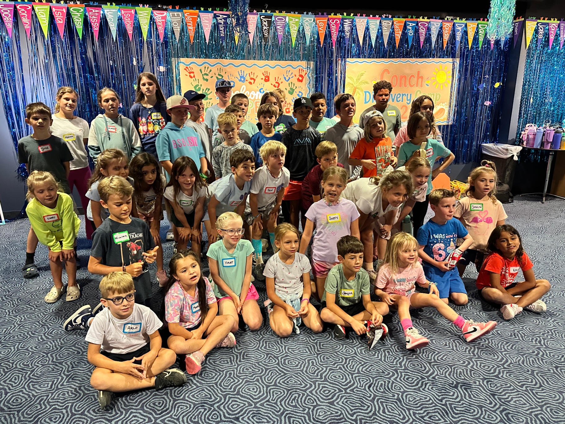 A large group of young campers sits together on a carpeted floor in front of a welcome backdrop, posing for a camp group photograph.