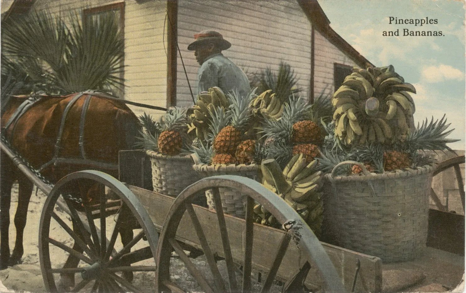 A horse-drawn wagon loaded with wicker baskets of pineapples and bunches of bananas in front of a clapboard building