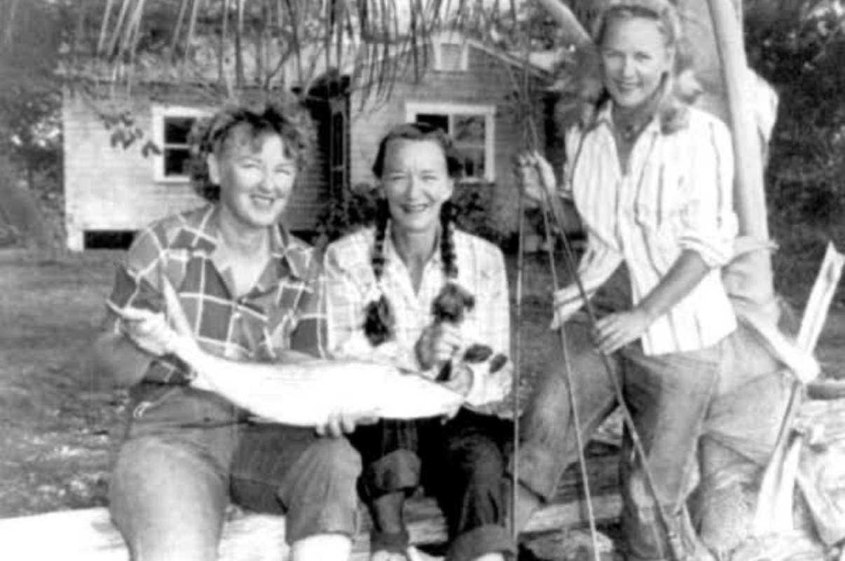 Black-and-white photograph of three smiling women holding a large bonefish in front of a wooden cottage, one leaning against a palm tree with fishing rods in hand