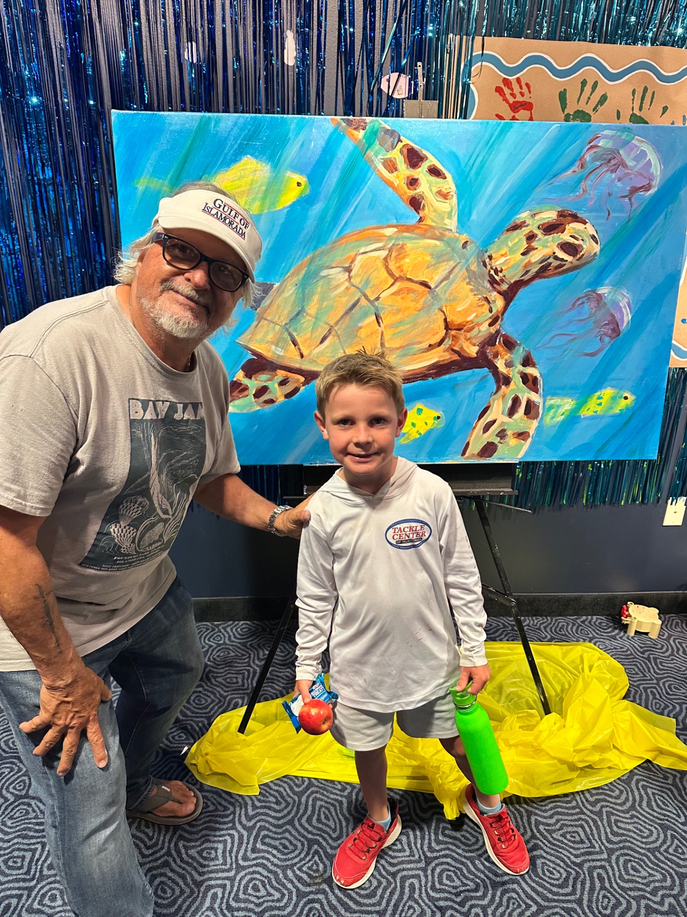 A smiling young camper stands beside Pasta the Painter in front of a painted sea turtle on an easel, holding an apple and a water bottle.