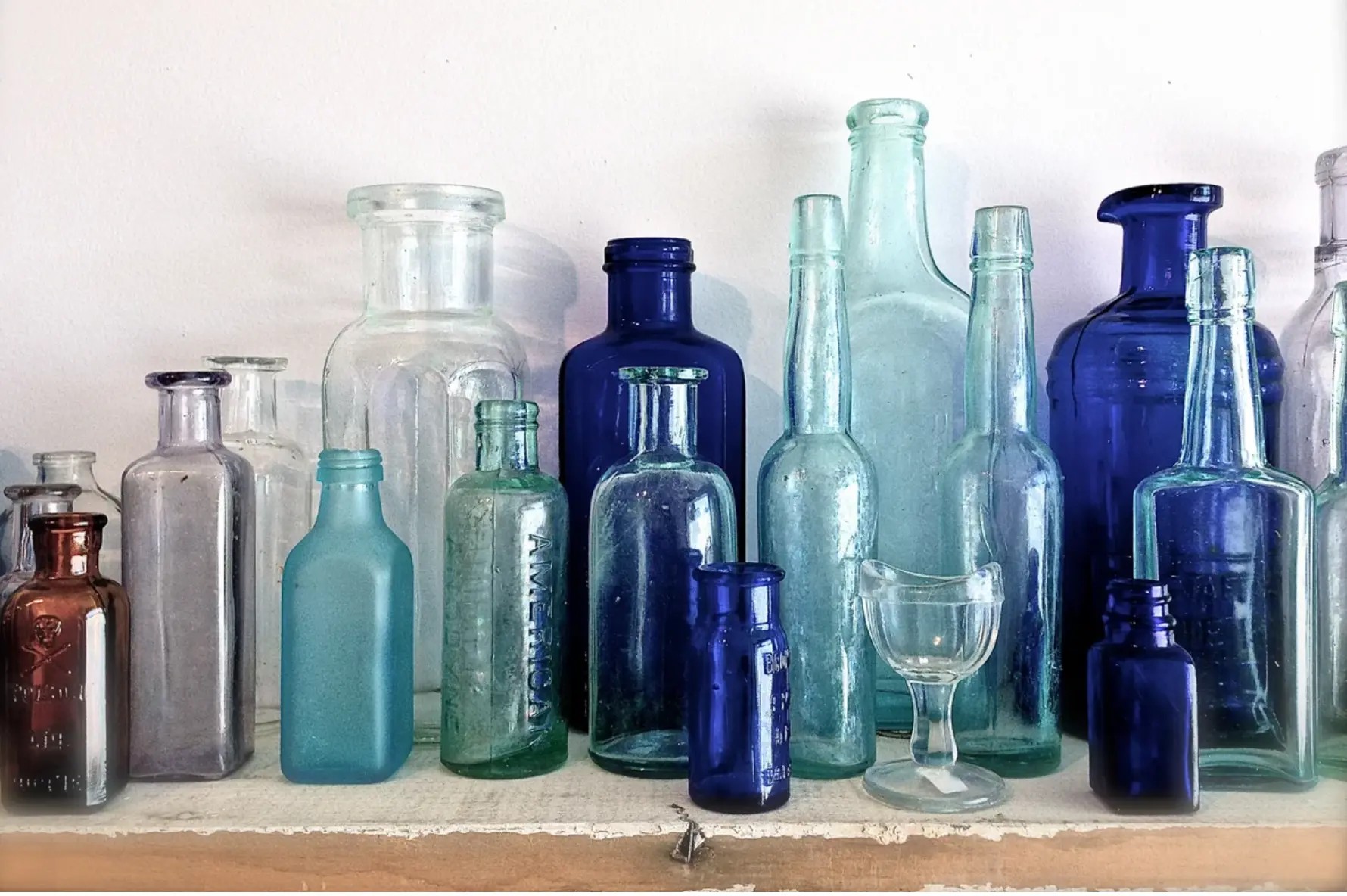 Collection of antique glass bottles and jars in cobalt blue, sea green, amber and pale lavender, including an amber bottle embossed with a skull and crossbones and the word POISON, arranged on a weathered white wooden shelf