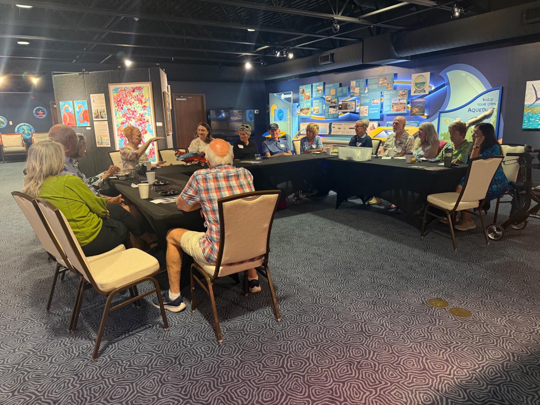 A curator leads conversation across a long table at Coffee with the Curator, with exhibit panels and the Build Your Own Aqueduct display in the background