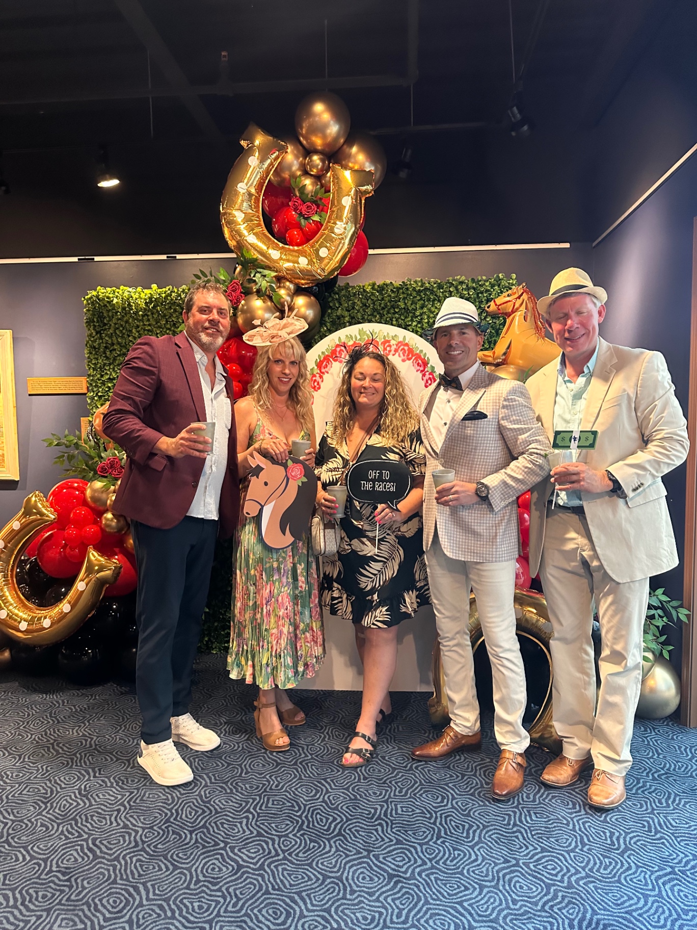 Five guests with festive hats pose at the Derby photo wall with gold horseshoe balloons