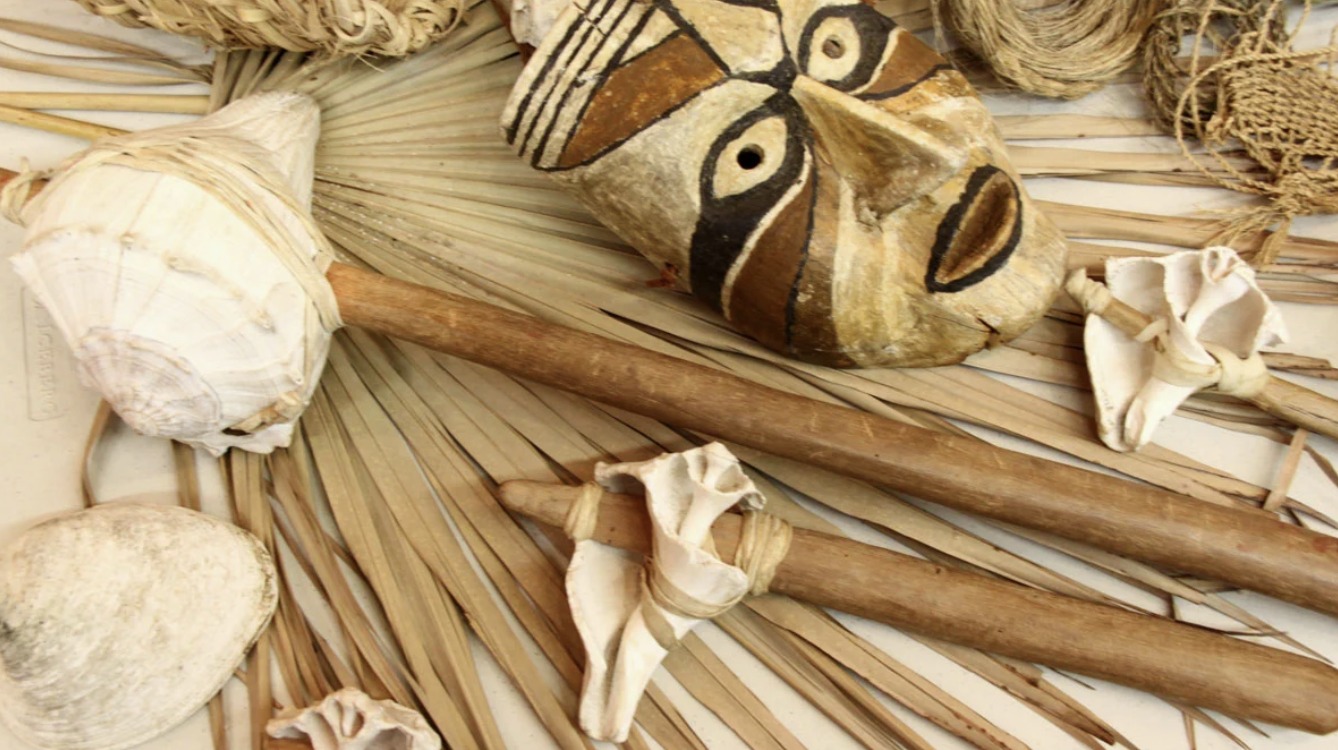 Overhead photograph of indigenous-style artifacts arranged on dried fan palm fronds: a shell-headed club lashed to a wooden handle, a carved and painted wooden mask, shell-bladed tools, and woven cordage