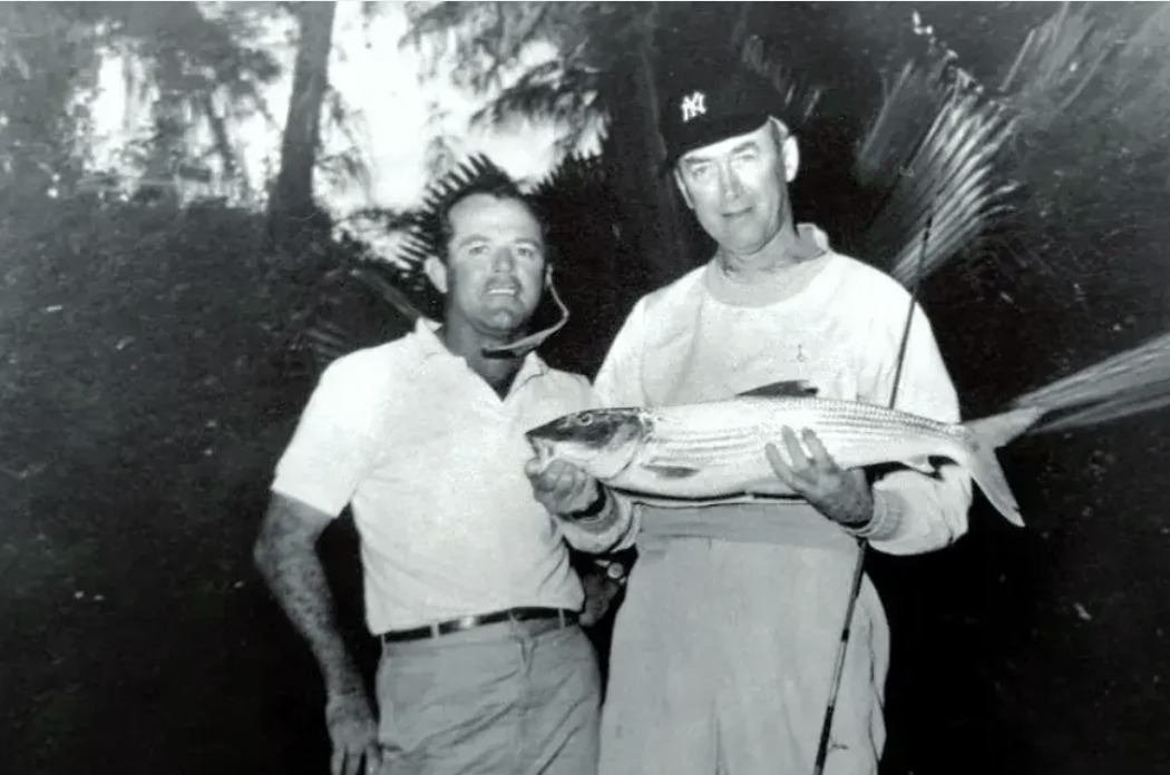 Black-and-white photograph of Captain Jimmie Albright at left and actor Jimmy Stewart at right holding a large bonefish between them, palm fronds behind and a fishing rod rising beside them