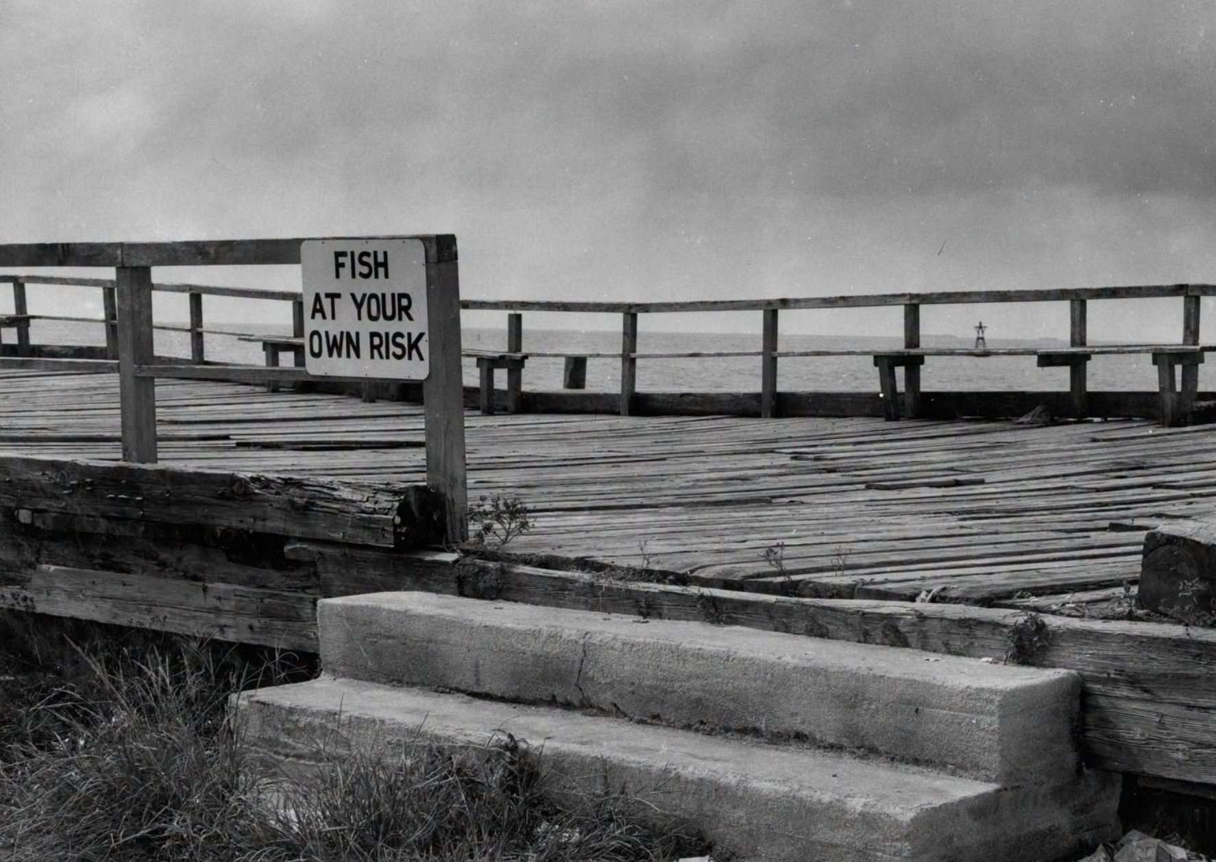Weathered wooden fishing pier over the water with a sign on the railing reading Fish at Your Own Risk.