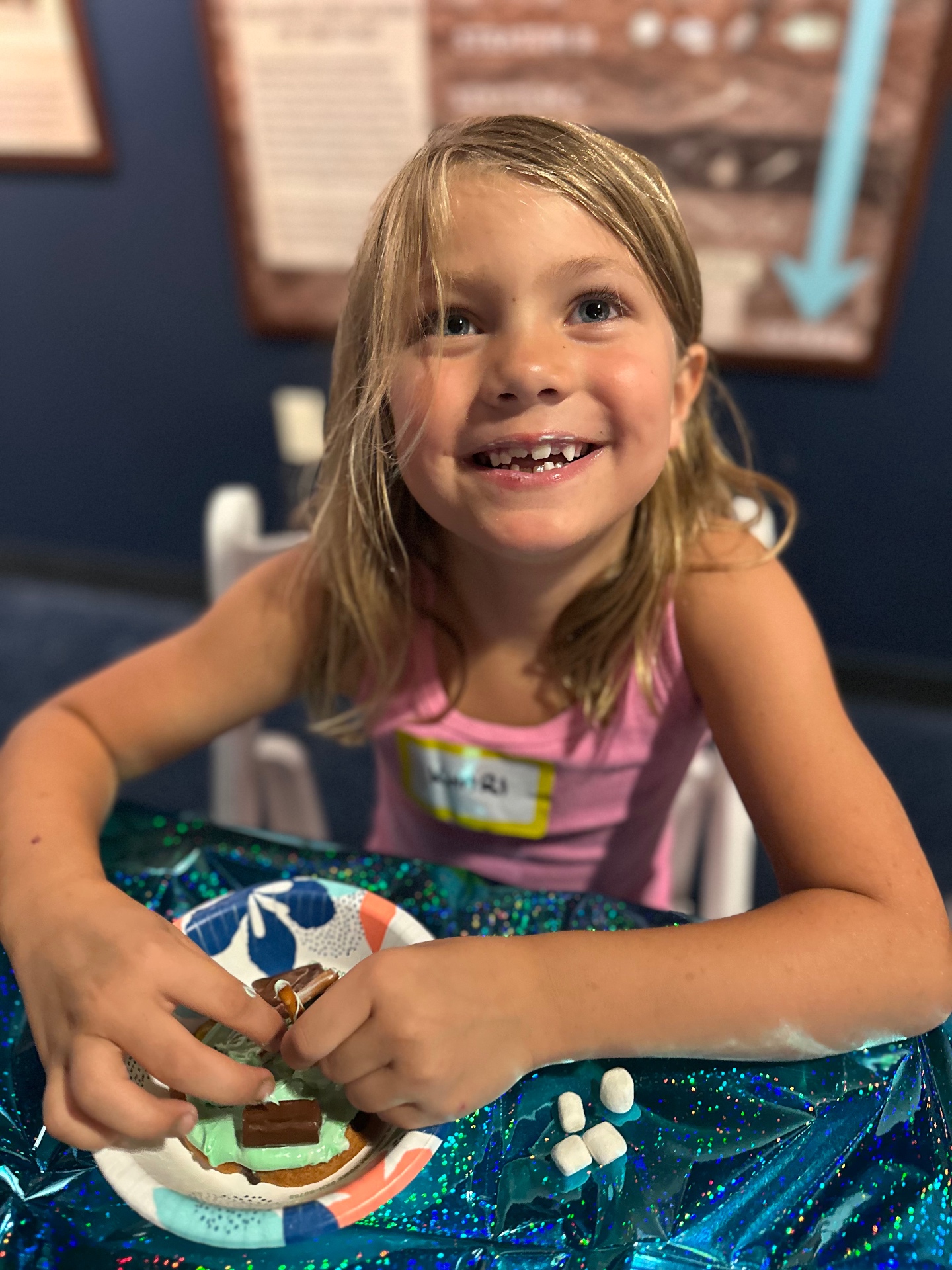 A young camper looks up and smiles while decorating a cookie with green frosting, candy and pretzels at a camp table inside the Discovery Center.