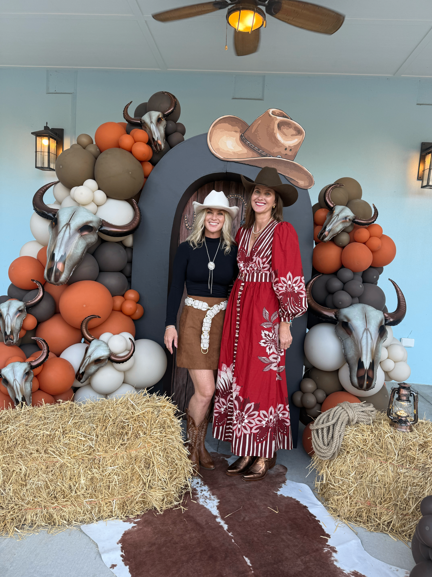 Two guests in Western wear pose at the Hoedown photo wall of balloons, longhorn skulls, and hay bales