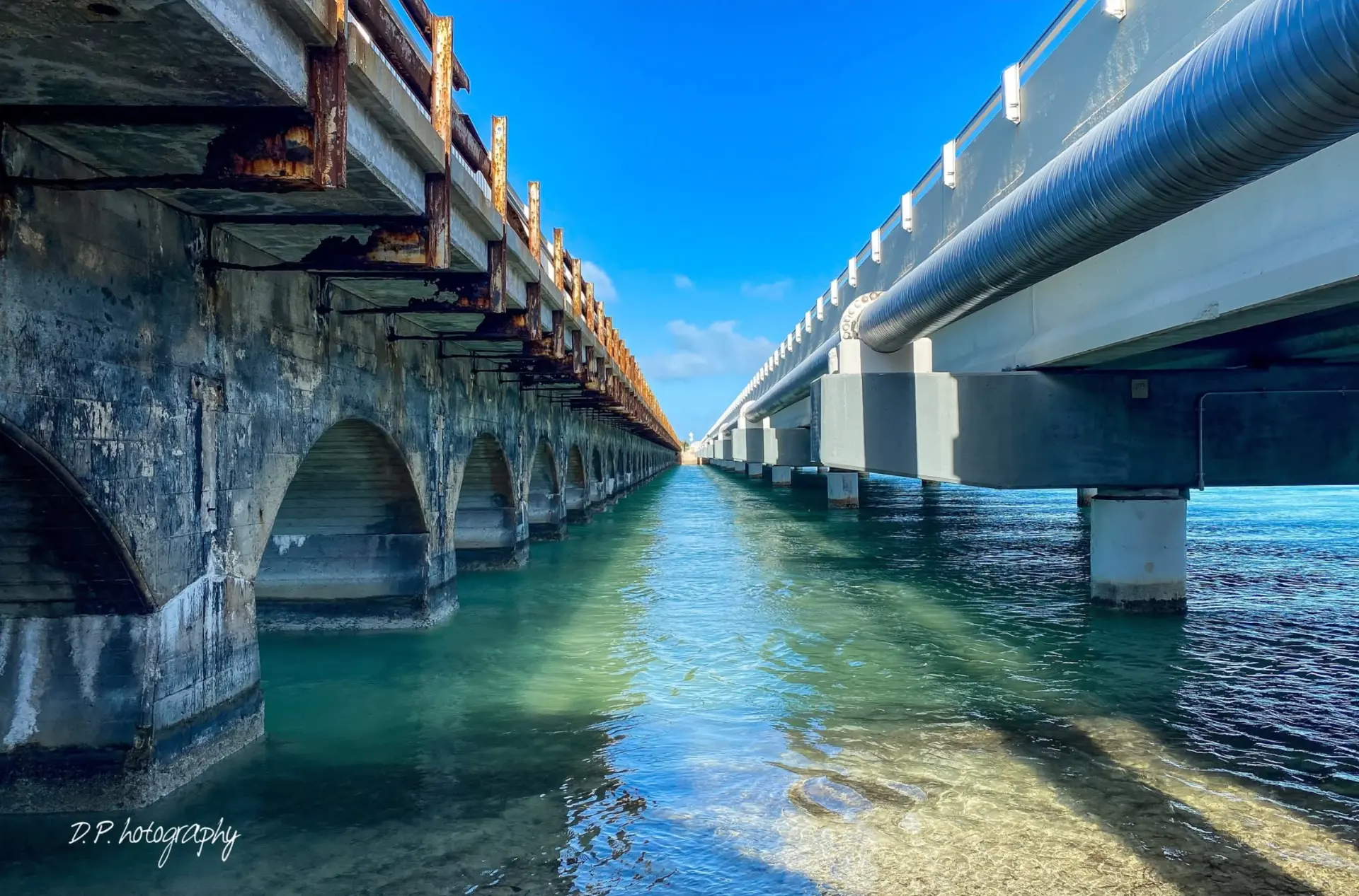 Photo looking down the channel beneath the Overseas Highway bridge, with the old railroad arches on one side and the modern bridge carrying the water pipeline on the other
