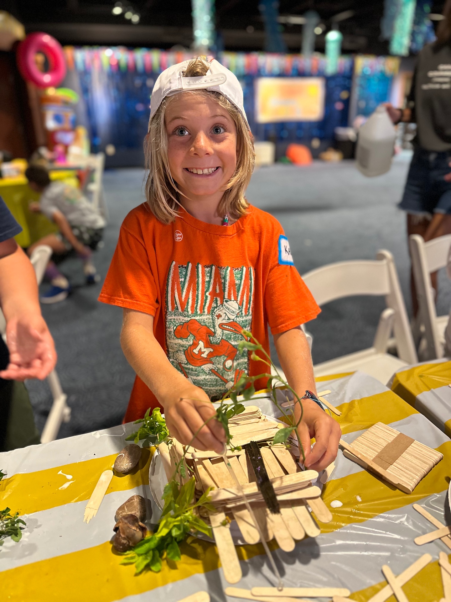A young camper in an orange shirt smiles broadly at the camera while holding up a nature craft built from wooden sticks, stones and plant clippings at a camp table.