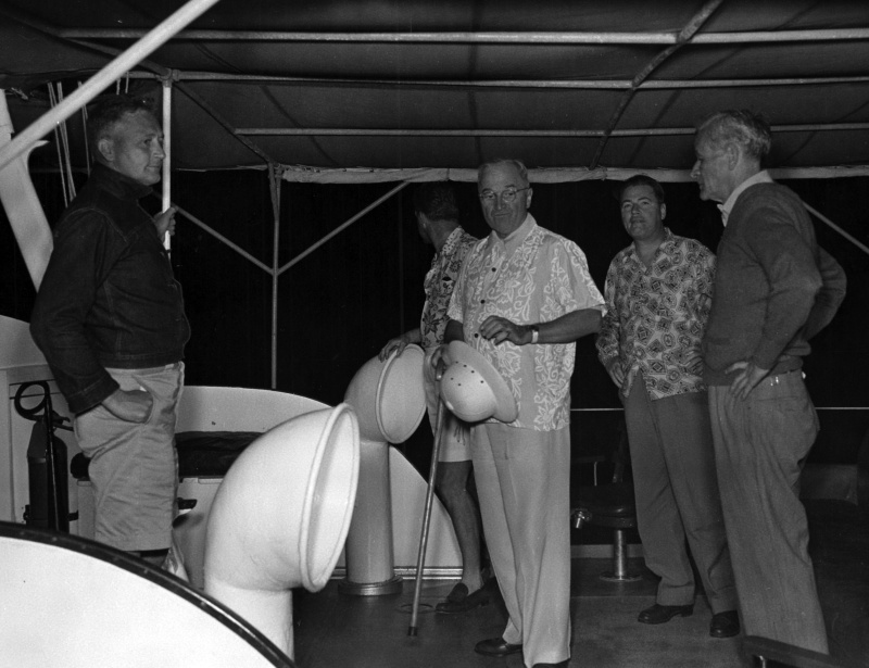 President Truman in a patterned shirt standing with several men under the awning of a boat in Key West, holding a cane and a pith helmet